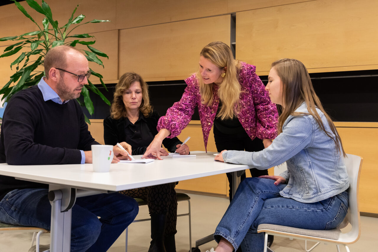 Een vrouwelijke trainer legt een concept uit aan een groep docenten tijdens een training, terwijl de docenten aandachtig luisteren.